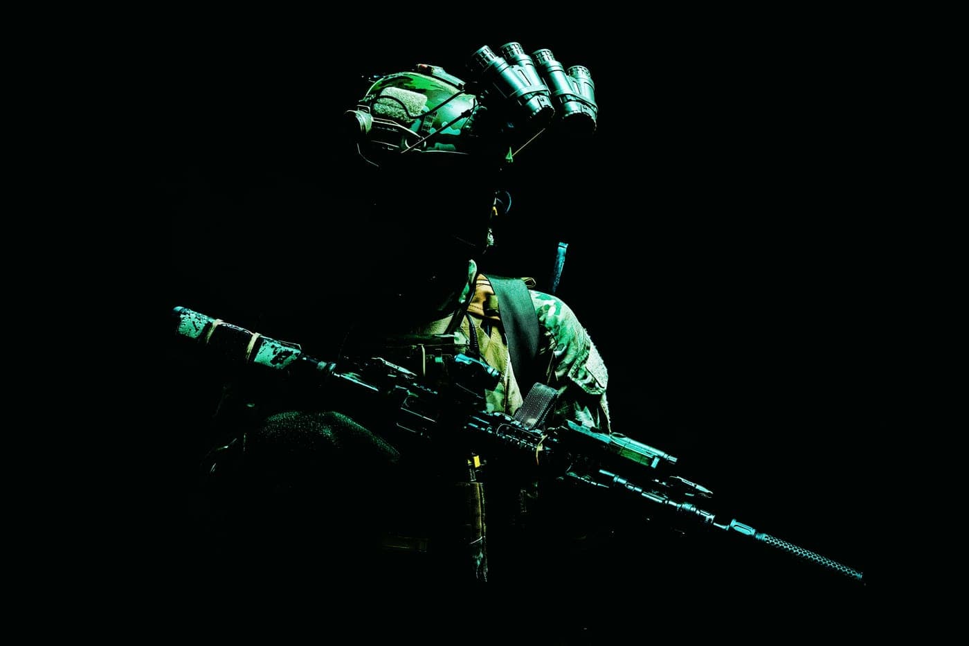 Service member wearing night-vision equipment, lit in green against darkness.